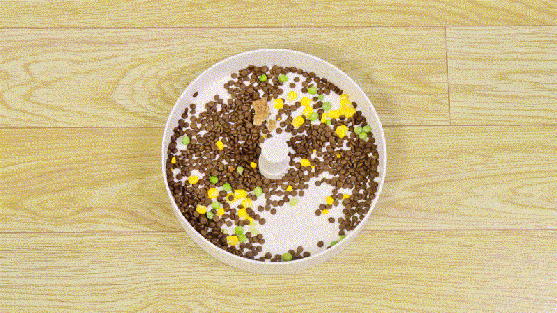 A round, white puzzle feeder filled with a mix of brown kibble and green, yellow, and orange pellet pieces is placed on a light wood floor.