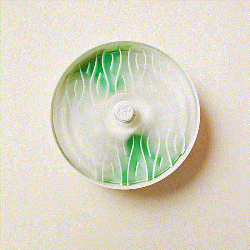 Round green and white device with spinning blades, mounted on a beige wall, designed as a feeder to slow down pets during mealtime.