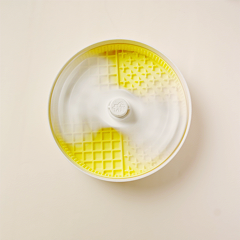 A spinning white and yellow kitchen salad spinner in motion viewed from above, resembling a puzzling whirlpool. The background is a plain beige surface.