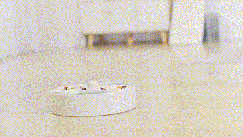 A sleek white pet food dispenser, resembling a modern dog bowl, sits gracefully on a light wooden floor, with a blurred white cabinet in the background.