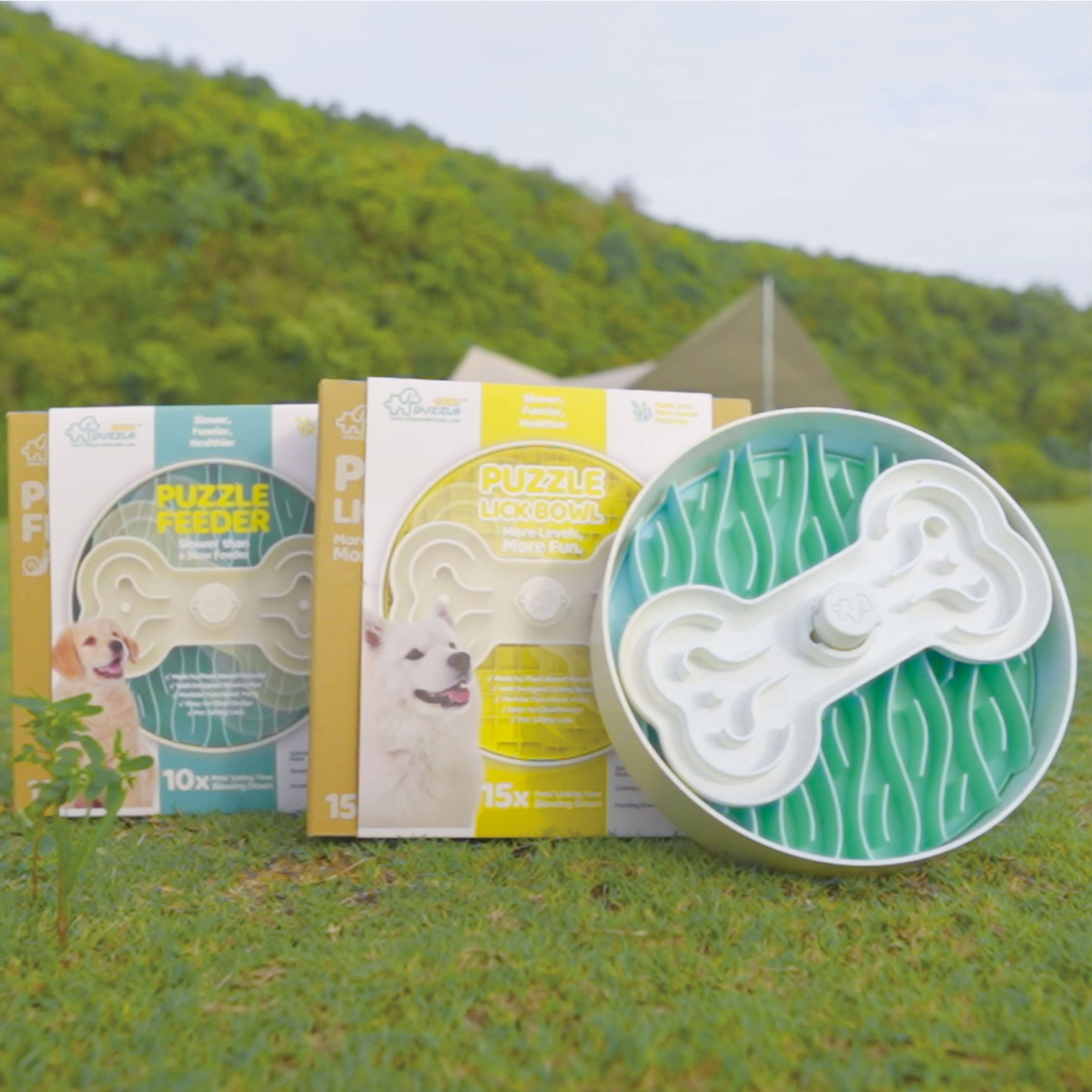 Three pet puzzle feeders are displayed outdoors. One feeder is green with a bone-shaped maze. The packaging for each feeder features images of dogs and product information. Trees and grass are in the background, creating a serene setting for these engaging puzzle bowls.