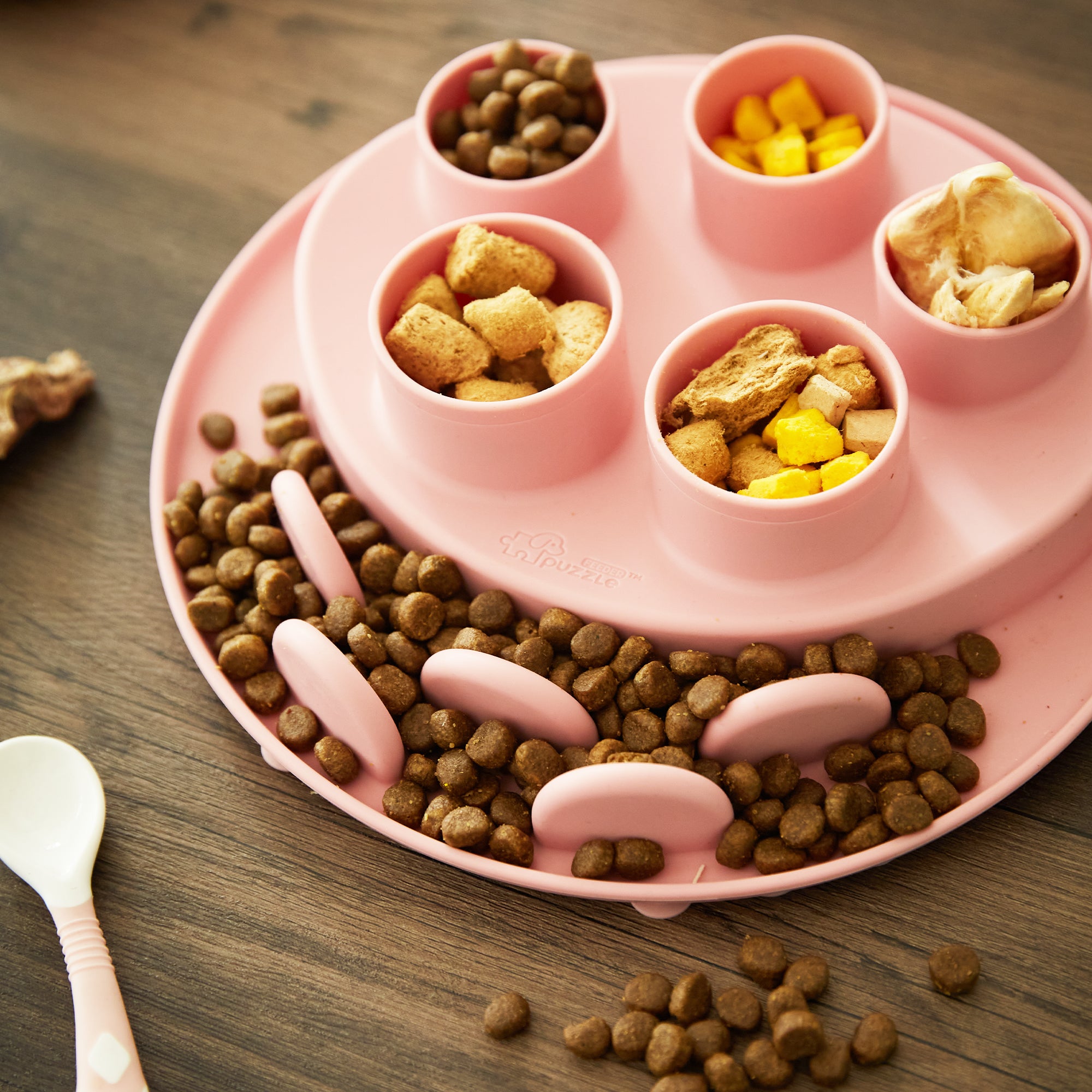 A pink slow feeder bowl filled with kibble, croutons, and diced mango. A white plastic spoon rests beside the bowl, designed to help your dog slow down while eating.