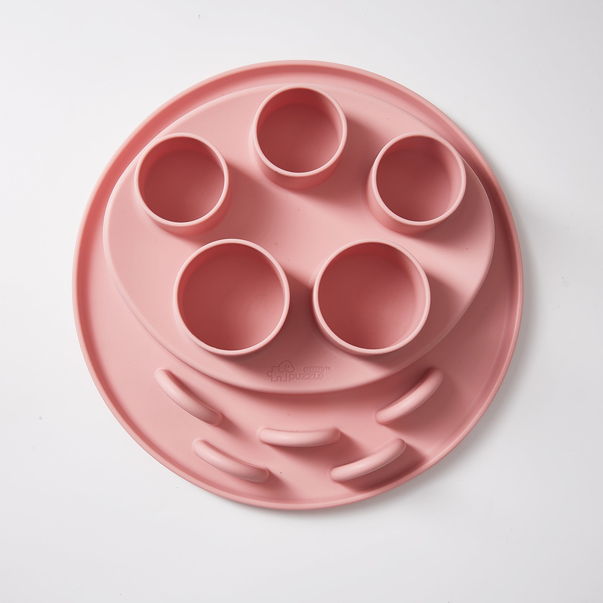 The Puzzle Digger by Puzzle Feeder™—designed in a coral-pink hue for cats—features five circular compartments and grooved pathways on a flat surface. Equipped with an antislip suction cup for stability, it enables your pet to engage their hunting instinct while exploring different sections.