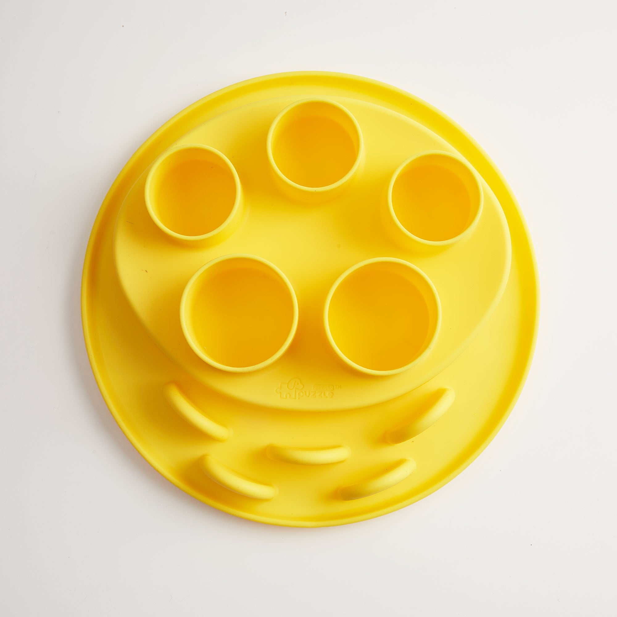 A wheat-yellow circular silicone tray, known as the Puzzle Digger by Puzzle Feeder™, features five cup holders and is made from safe materials, sitting on a white background.
