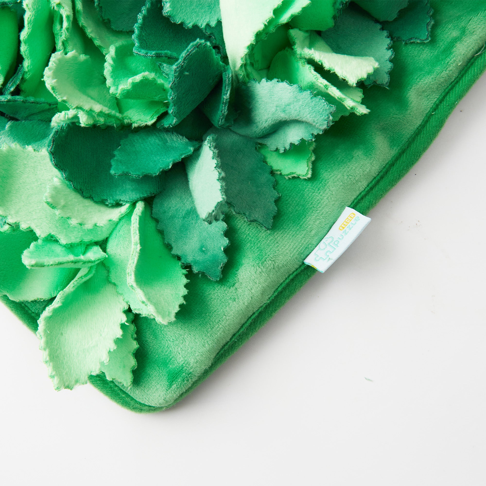 A close-up of the Puzzle Feeder™ Dog Puzzle Grass Sniffle Mat showcases its green fabric with layered, leaf-like textures that resemble realistic grass, making it ideal for sensory stimulation. A small tag in the corner adds an extra touch of detail.