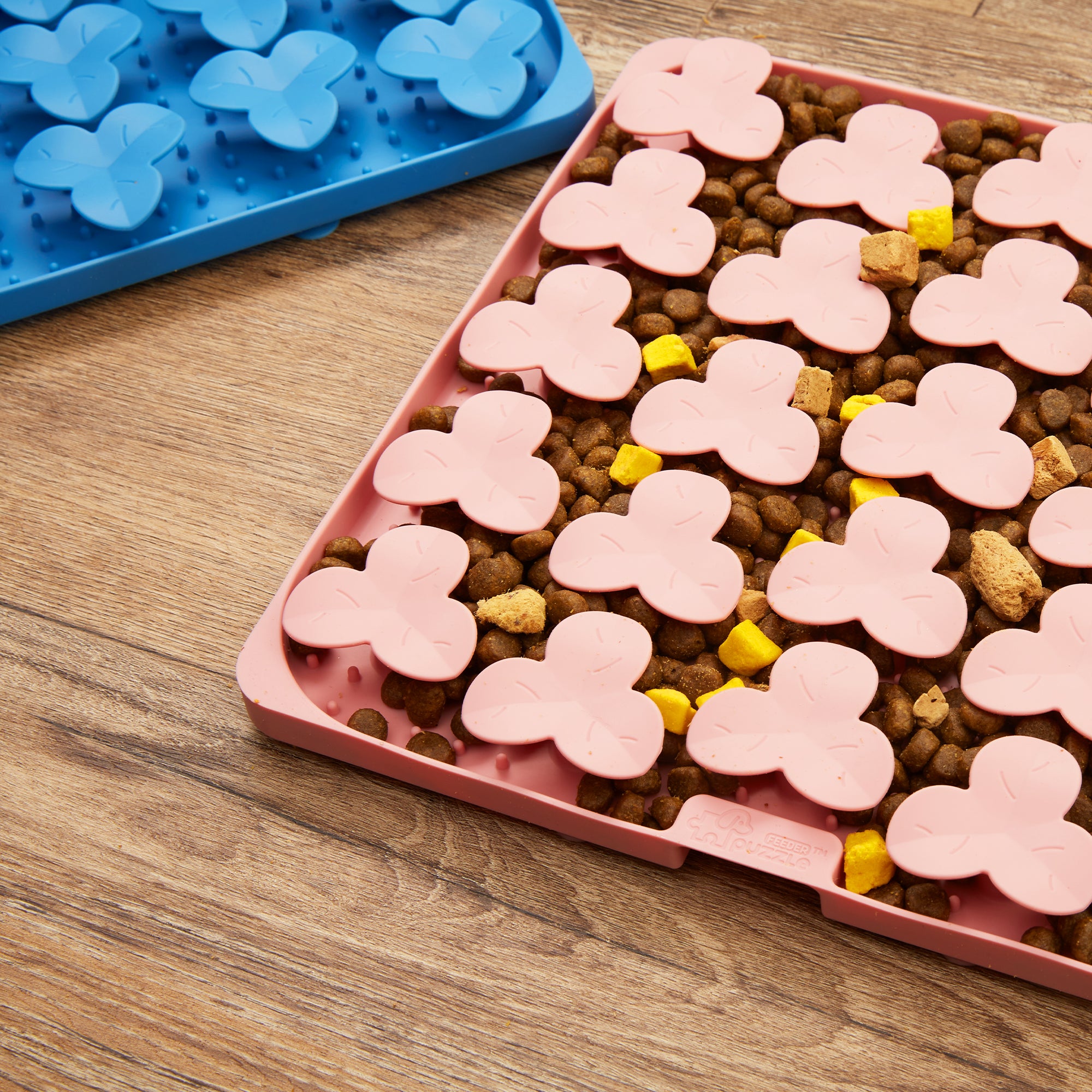 Two trays with butterfly-shaped inserts are placed on a wooden floor; one tray is blue and empty, the other is a pink puzzle feeder filled with brown pellets and a few yellow cubed pieces to help slow down feeding.
