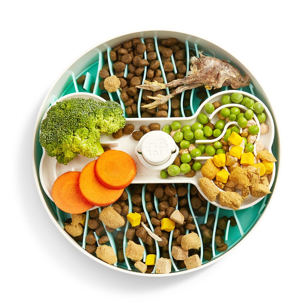 The Puzzle Feeder™ Swirl - Slower than a Slow Feeder - For Dogs (Purple) by Puzzle Feeder™ encourages healthy eating habits when filled with dry pet food, broccoli, carrot slices, peas, yellow cubes, and a small bone arranged around a white divider on a blue base.