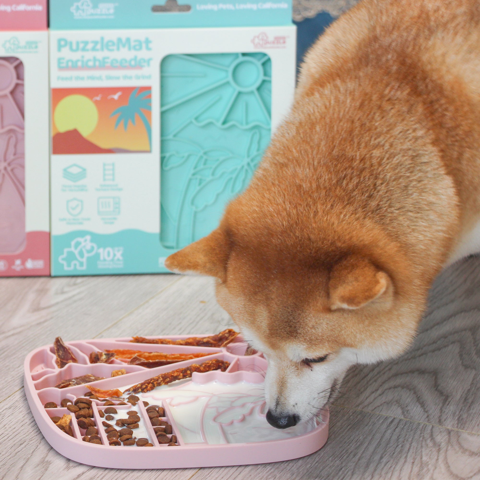 A Shiba Inu dog enjoys a meal from a pink Puzzle Feeder™ California Themed Lick Mat, promoting slow eating for healthy digestion. In the background, teal and pink packaging highlights its California-inspired design.