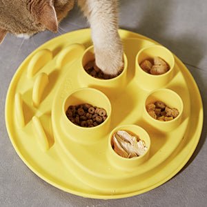 A cat paws at food in the Puzzle Feeder™ Dog Puzzle Digger - For Hunting Instinct (Wheat-Yellow), designed from non-toxic materials with multiple compartments to mimic hunting instincts, on a gray surface.