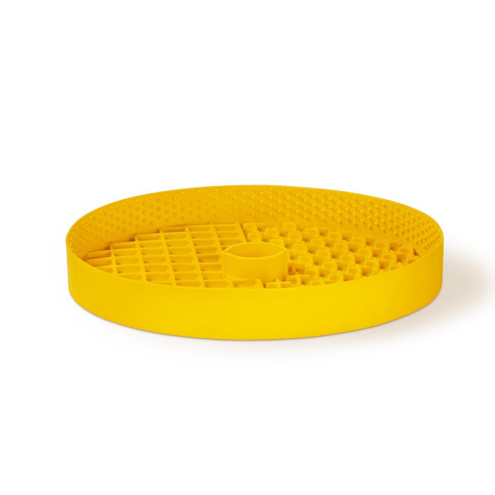 The Dog Licking Mat Accessory by Puzzle Feeder™ is a yellow, oval-shaped rubber mat featuring a textured surface and a round raised center, specifically designed as a slow feeder dog bowl to keep your pet engaged.