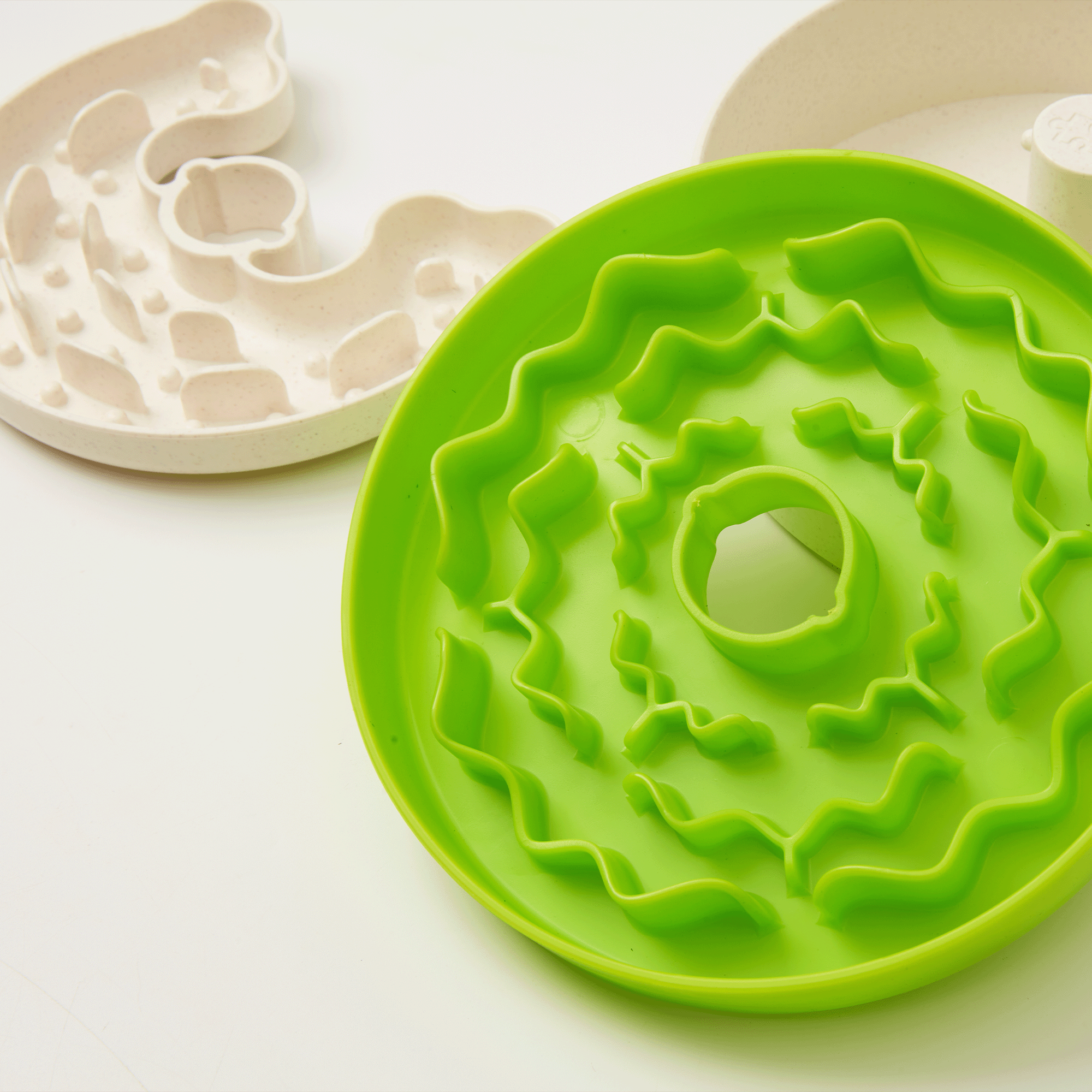 Two Dog Puzzle Feeder™ Lite bowls from Puzzle Feeder™ are shown, one green with a spiral design and one white with a maze configuration. These bowls are designed to slow down pet eating and offer versatile feeding compatibility for your furry friend.