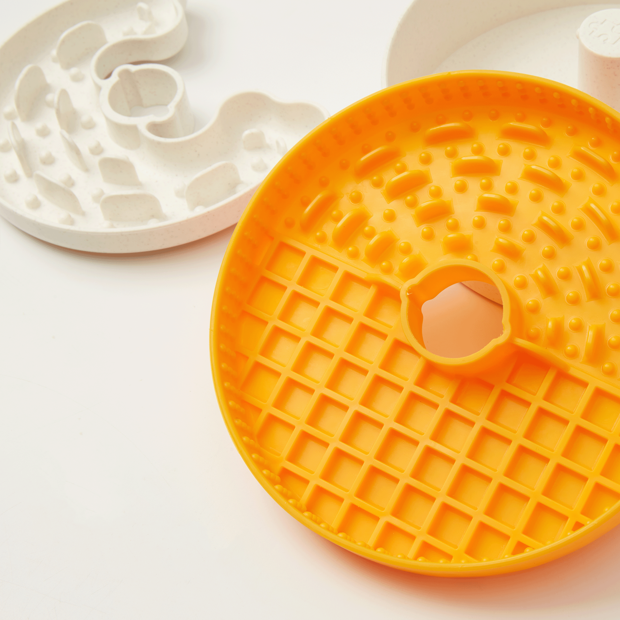 An orange circular Dog Puzzle Lick Bowl Lite by Puzzle Feeder™ sits prominently with a grid pattern in the center, while a white maze-shaped toy rests nearby, both placed on a white surface. This engaging setup doubles as an interactive puzzle feeder, providing mental stimulation for small and medium breeds of dogs during licking training.