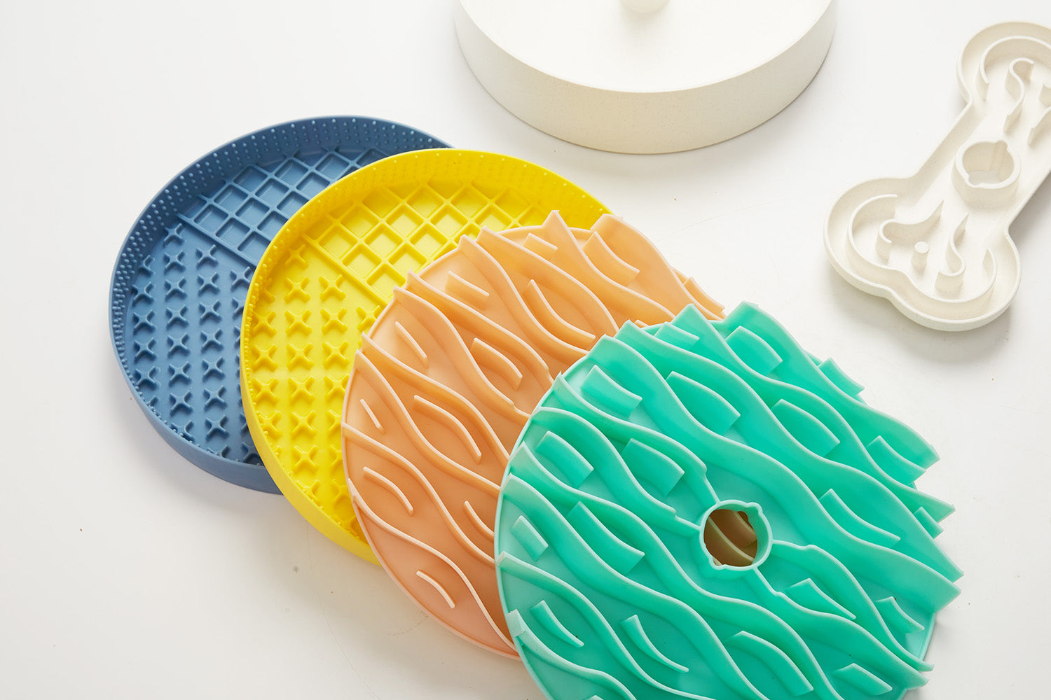 Four textured rubber mats in blue, yellow, orange, and teal are arranged on a white surface alongside a sleek dog bowl and a bone-shaped accessory.