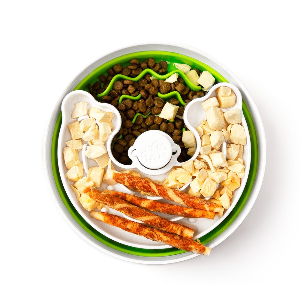 The Puzzle Feeder™ Junior, crafted by Puzzle Feeder™, is a white and green divided pet food bowl designed for small dogs and puppies to encourage healthy eating habits. It holds kibble, diced chicken, green garnish, and three chicken sticks while serving as a puzzle feeder for an engaging mealtime experience.