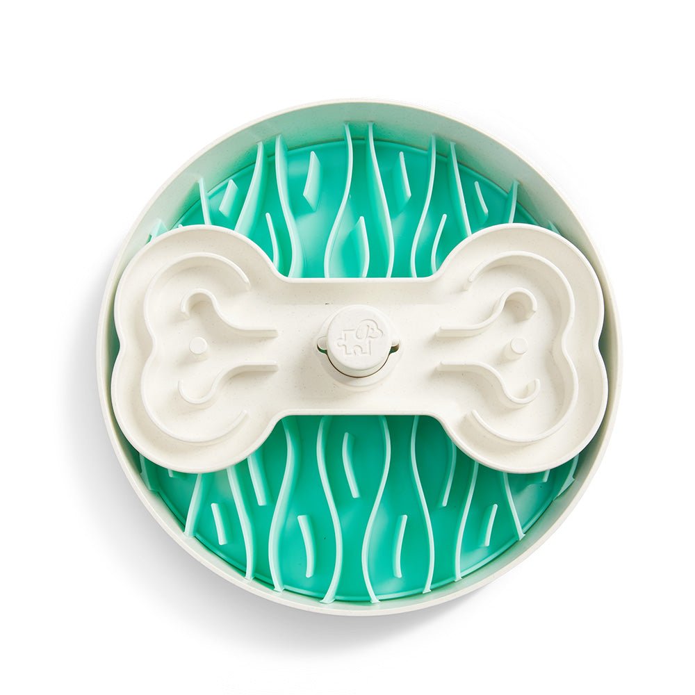 The Puzzle Feeder™ Swirl by Puzzle Feeder™ is a circular dog bowl featuring a turquoise interior and a white, bone-shaped lid with grooves, designed to promote healthy eating habits as a slow feeder.