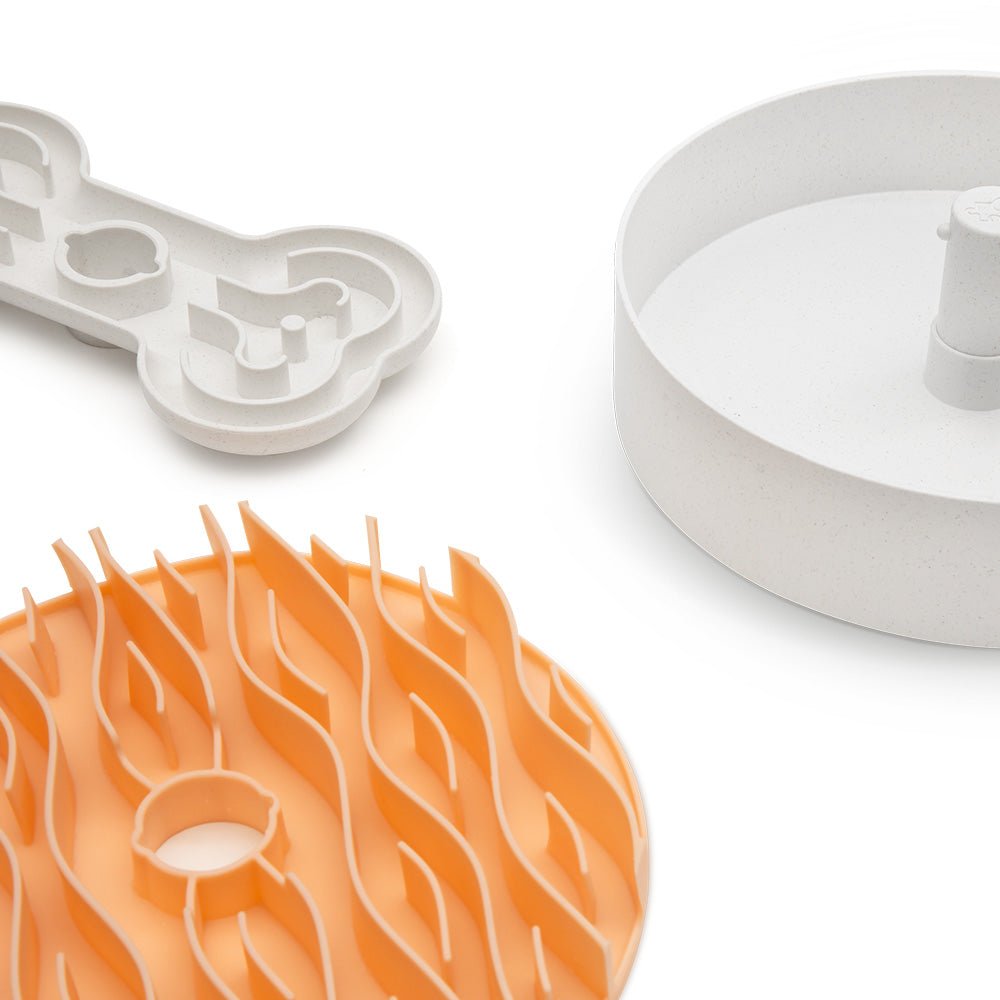 A collection of Puzzle Feeder™ components, featuring a circular mold in white, a wavy grid in orange, and a bone-shaped piece reminiscent of elements from the Puzzle Feeder™ Wave for Dogs (Pink), is artfully arranged on a white background.