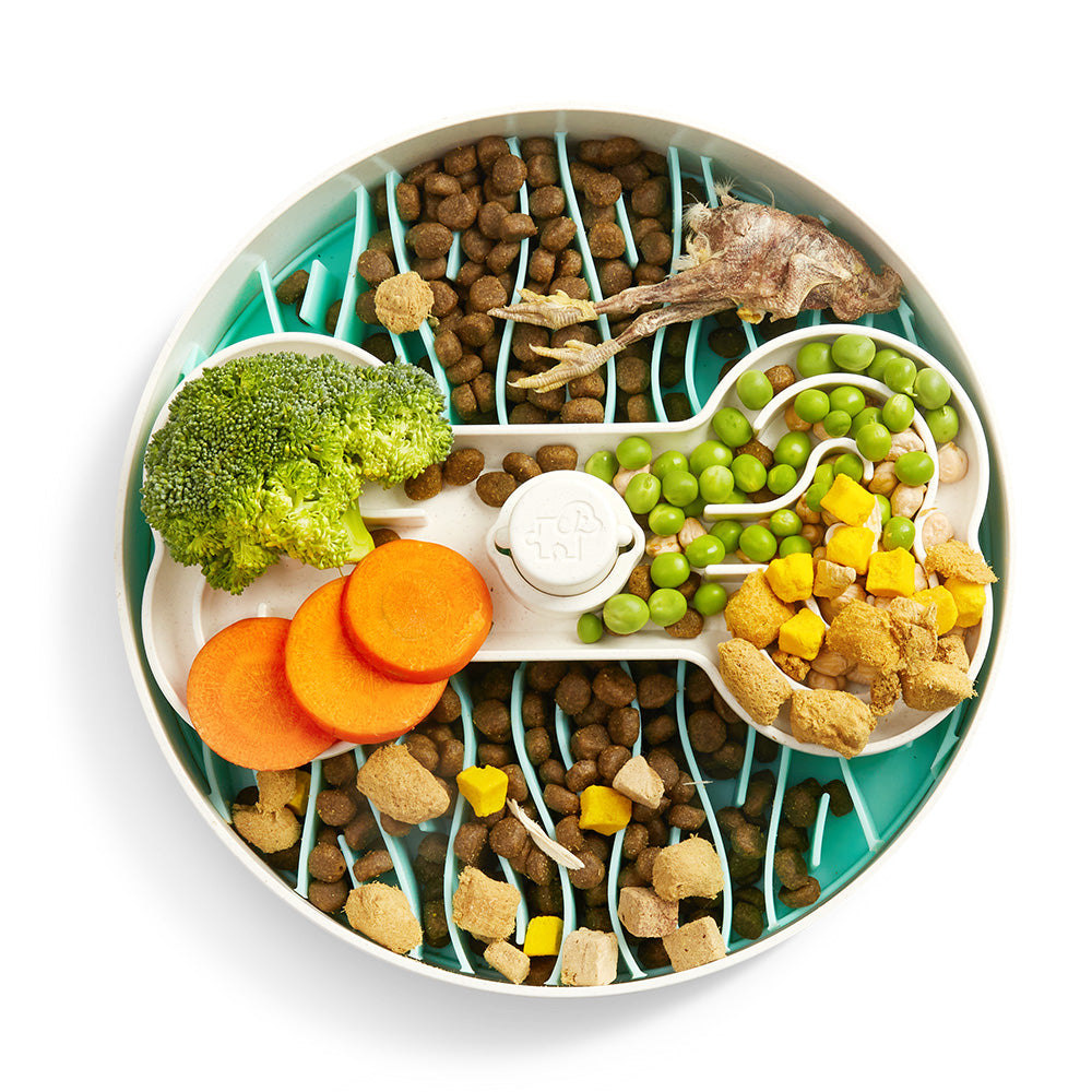 The Puzzle Feeder Premium Double Kit from Puzzle Feeder™ is filled with broccoli, carrot slices, peas, kibble, and a chicken wing as it sits on a white and teal patterned surface. This arrangement not only promotes healthy digestion but also provides an entertaining puzzle feeding experience for your pet.