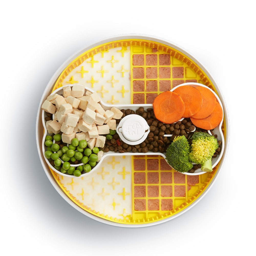 The Puzzle Licker Maze - Triple Textures, Triple Fun (Yellow) by Puzzle Feeder™ features a bone-shaped puzzle center that holds diced chicken, peas, kibble, broccoli, and carrot slices on a textured yellow surface designed to promote dog dental health.