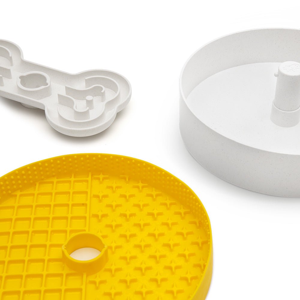 The Puzzle Licker Maze by Puzzle Feeder™ features three plastic components in white and yellow colors, resembling slow feeder dog bowls. It includes a bone-shaped piece, a circular piece with a central post, and a yellow grid-patterned round piece designed for enhancing dog dental health.