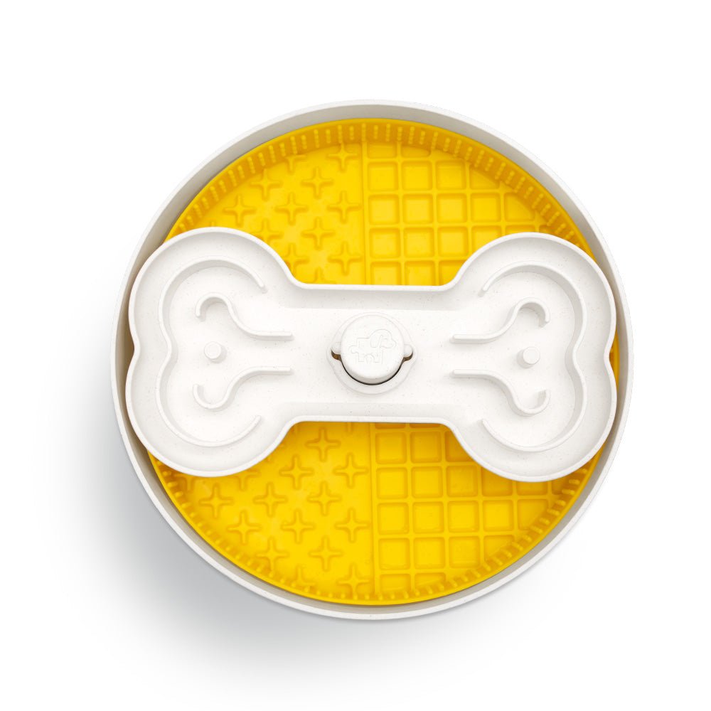 The Puzzle Licker Maze - Triple Textures, Triple Fun (Yellow) by Puzzle Feeder™ is a yellow, textured feeder created to enhance dog dental health, featuring a distinctive white bone-shaped cover in the center.