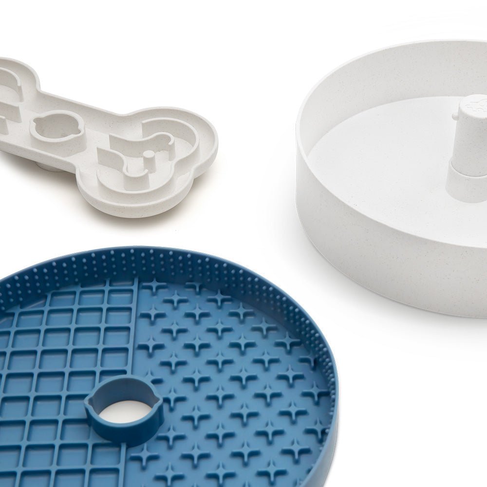 A bone-shaped tray, the Puzzle Licker Maze from Puzzle Feeder™ featuring a textured blue circular design similar to puzzle feeder bowls, and a plain white circular tray with a central post are arranged on a white surface.