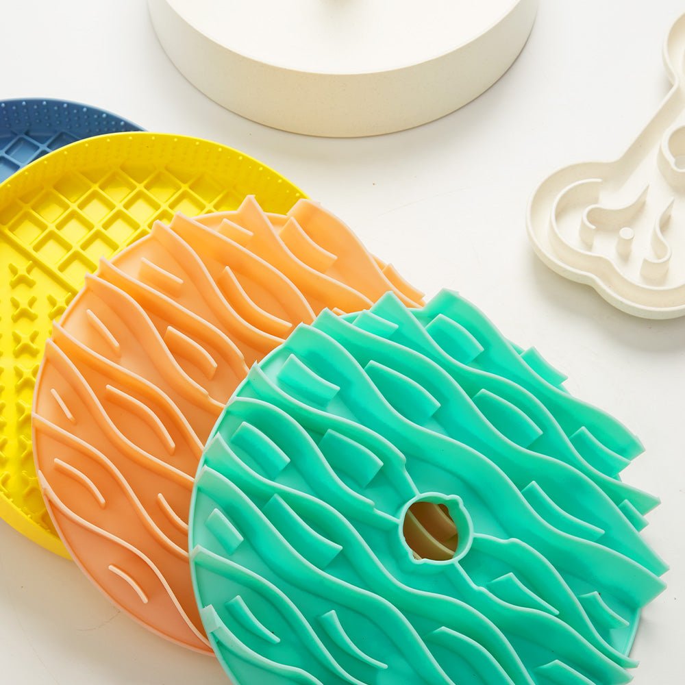 On a white surface, four vibrant silicone mats from Puzzle Feeder™ (Dog Slowing Mat Accessory) in yellow, orange, and teal are displayed. These mats feature circular and abstract wavy patterns, designed to engage pets during mealtime with their unique puzzle feeder functionality.