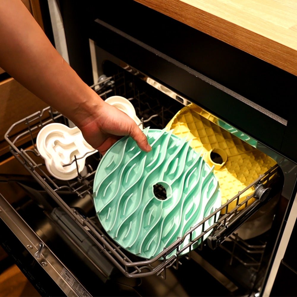 A hand places a turquoise Dog Slowing Mat Accessory by Puzzle Feeder™—designed with a pet-friendly aesthetic—into a dishwasher, alongside other molds in white and yellow.